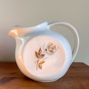 May include: White ceramic pitcher with a brown floral design featuring a single rose. The pitcher has a rounded body and a handle.