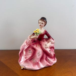 May include: A porcelain figurine of a woman in a long, pink dress with a dark red bodice. She holds a bouquet of yellow flowers. The dress has gold accents and a floral pattern. The figurine is on a wooden surface.