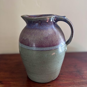 May include: A small, hand-thrown ceramic pitcher with a light green glaze and a purple and blue glaze on the top. The pitcher has a rounded body and a handle.