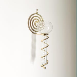 May include: A wall-mounted decorative glass vase with a gold-colored metal spiral design. The vase has a clear glass tube and a round glass bowl. The metal spiral is attached to a circular design for hanging.