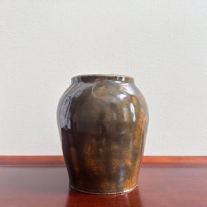 May include: A handcrafted, brown ceramic vase with a glossy finish. The vase has a rounded body and a slightly flared rim. The color transitions from dark brown to a lighter, reddish-brown near the base. The vase is displayed on a wooden surface.