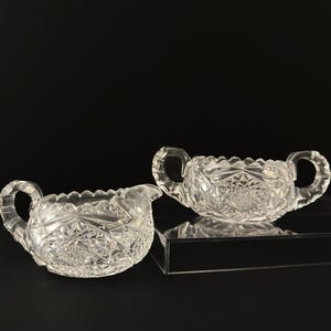 May include: A set of two clear glass sugar bowls with cut glass designs. The bowls have two handles each and are sitting on a clear acrylic surface.