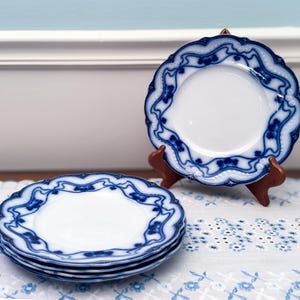 May include: A stack of four white porcelain plates with a blue floral design and scalloped edges. One plate is displayed on a wooden stand. The plates are on a white tablecloth with a blue floral pattern.