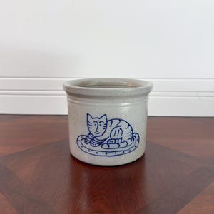 May include: A cylindrical stoneware crock with a speckled light gray finish. The front features a hand-painted blue line drawing of a cat resting on a patterned mat. The vessel has a smooth, glazed surface and a simple, straight-sided design.