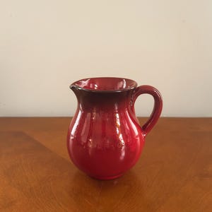May include: A glossy red ceramic pitcher with a dark brown rim and handle. The pitcher has a rounded body and a spout for pouring. The surface reflects light, highlighting its smooth texture. A classic kitchenware item.