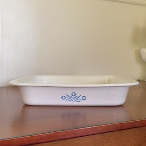May include: A rectangular white ceramic baking dish with rounded corners and a wide rim. The exterior features a centered blue floral pattern consisting of a small bouquet of stylized flowers and leaves. The dish has a smooth, glossy finish.