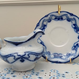 May include: A white and blue porcelain creamer and saucer set. The creamer has a handle and spout, and the saucer is round. Both pieces feature a blue floral design around the edges. The set is displayed on a white and blue patterned cloth.