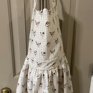 May include: A cream-colored apron with a gingham pattern and a repeating design of chickens in various poses. The apron has a ruffled skirt and a bow at the neck. The apron is hanging on a door.