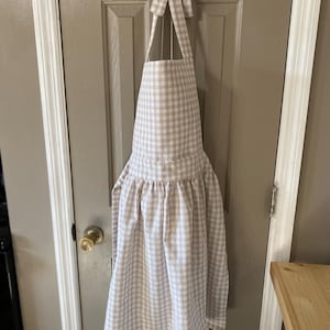 May include: A beige and white gingham apron with a ruffled hem and a bow at the neck.