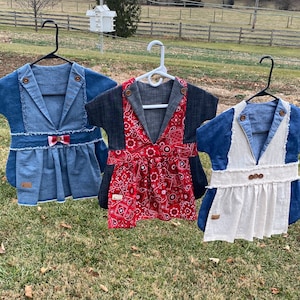 May include: Three denim and fabric dresses hanging on hangers. The dresses have a blazer-style top with different fabric patterns, including denim, red paisley, and white linen. Each dress has wooden buttons and a small tag.
