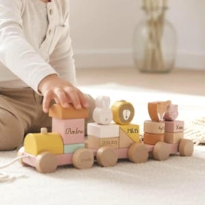 Puede incluir: Un tren de juguete de madera en colores pastel con varias formas de bloques, incluyendo un conejito y un tronco. El tren tiene el nombre "Ambre" en un bloque y otros bloques con fechas y números. El tren está sobre una superficie clara.