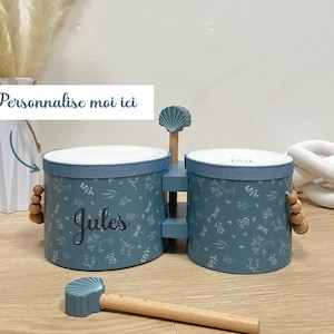 Könnte beinhalten: Ein blaues Holzspielzeug-Schlagzeug mit einer weißen Oberseite und einem Blumenmuster. Das Schlagzeug hat einen Holzstock mit einer blauen Muschel am Ende. Das Schlagzeug ist mit dem Namen "Jules" personalisiert.