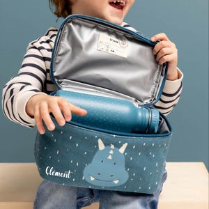 Puede incluir: Una bolsa de almuerzo azul con un diseño de dinosaurio y el nombre "Clément" en blanco. La bolsa está abierta, revelando una botella de agua azul con lunares blancos. El interior de la bolsa es plateado.