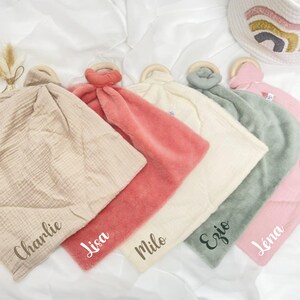 May include: Five baby security blankets with wooden rings. The blankets are in different colors and textures, including beige, pink, cream, green, and light pink. Each blanket has a name embroidered on it: Charlie, Lisa, Milo, Ezio, and Lena.