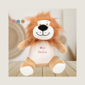 Può includere: Un leone di peluche marrone con la faccia bianca e un disegno ricamato rosa e bianco sul petto. Il disegno include due cuori e il nome "Pauline".