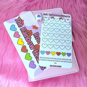 May include: A pink game set featuring the text "Roll The Rainbow Hearts" and colourful heart illustrations. Includes a die and cards with heart outlines for colouring. The set is on a pink surface.
