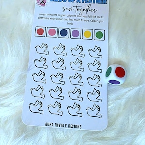 May include: A white card with the text "BIRDS OF A FEATHER save together" and a dice. The card has a grid of bird outlines and colour-coded boxes. A multi-coloured die is next to the card. The text "AURA ROYALE DESIGNS" is at the bottom.