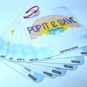 May include: A set of square cards held together by a pink ring, with the text "POP IT & SAVE" and a rainbow design. The cards have a watercolour background and display savings amounts. The brand name "Aura Royale Designs" is also visible.