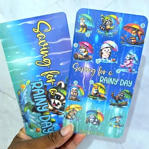May include: Two rectangular savings trackers with a blue and green gradient background featuring rain patterns. The text reads Saving for a RAINY DAY. One tracker shows a raccoon, while the other displays various animals with umbrellas.