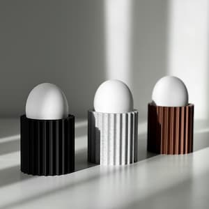 Può includere: Tre oggetti bianchi a forma di uovo sono esposti in tre porta uova di diversi colori. I porta uova sono realizzati in un materiale strutturato e hanno un design a coste. I porta uova sono nero, grigio e marrone.