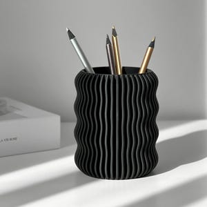 Pode incluir: Porta-canetas preto ondulado com quatro canetas dentro. As canetas são prateadas, douradas e pretas.