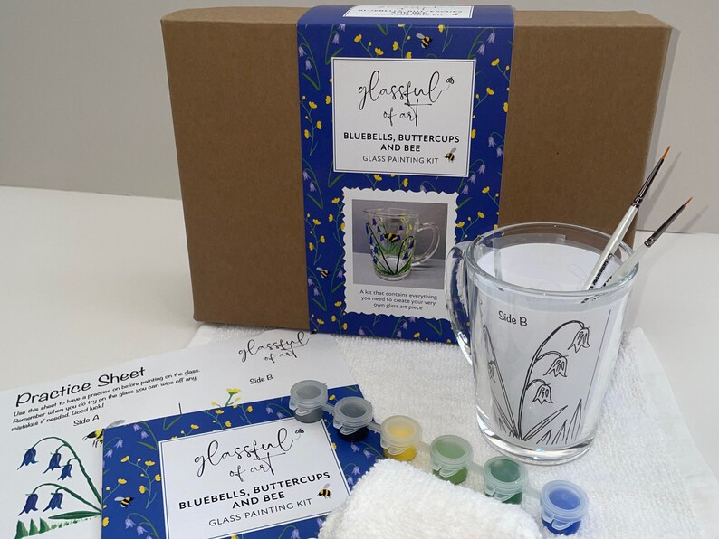May include: A glass painting kit featuring a clear glass mug with a bluebell, buttercup, and bee design. The kit includes paints, brushes, and a practice sheet. The box is blue with a floral pattern and the text "glassful of art BLUEBELLS, BUTTERCUPS AND BEE GLASS PAINTING KIT".