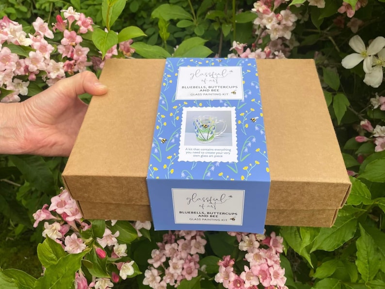 May include: A brown box with a blue and white label. The label has a picture of a mug with bluebells, buttercups, and a bee. The text on the label reads "glassful of art BLUEBELLS, BUTTERCUPS AND BEE GLASS PAINTING KIT".