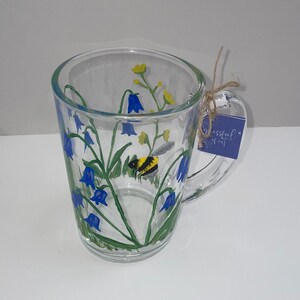 May include: A clear glass mug with a painted design of bluebells, green leaves, and a yellow and black bumblebee. The mug has a handle and a blue tag that says "Blissful"