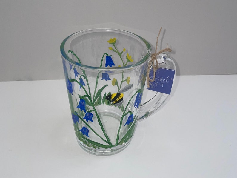 May include: A clear glass mug with a painted design of bluebells, green leaves, and a yellow and black bumblebee. The mug has a handle and a blue tag that says "Blissful"