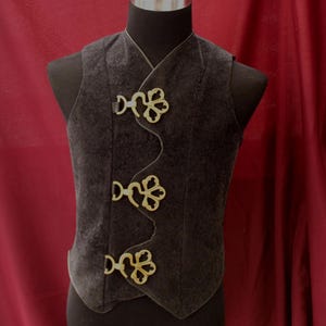 Peut inclure: Un gilet gris foncé avec trois fermoirs dorés ornés. Le gilet est présenté sur un mannequin noir sur fond rouge. Les fermoirs ont un motif floral.