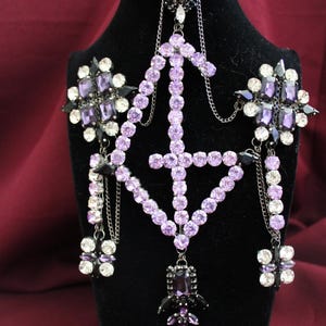 Può includere: Una collana ornata con un design centrale di cristalli color lavanda. La collana presenta gruppi di cristalli neri e trasparenti, collegati da delicate catene. È esposta su una testa di manichino nera su sfondo bordeaux.