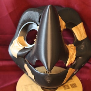 Könnte beinhalten: Eine schwarze und braune Fellmaske mit einer großen, spitzen Nase. Die Maske besteht aus einem glatten, harten Material und hat zwei große Augenlöcher. Die Maske ist teilweise mit braunem Klebeband bedeckt.