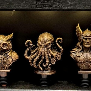 Pode incluir: Três rolhas de garrafa com acabamento metálico na cor bronze. A rolha da esquerda apresenta uma coruja com detalhes de engrenagens, a central um polvo com tentáculos e engrenagens, e a da direita um busto de guerreiro com capacete alado.