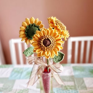 Könnte beinhalten: Ein Strauß gehäkelter Sonnenblumen in einer durchsichtigen Glasvase mit einem braunen Band. Die Sonnenblumen sind gelb mit grünen Blättern und braunen Zentren. Die Vase steht auf einem Tisch mit einer grün-weiß karierten Tischdecke. Der Text "Color Crochet BG" ist unten im Bild sichtbar.