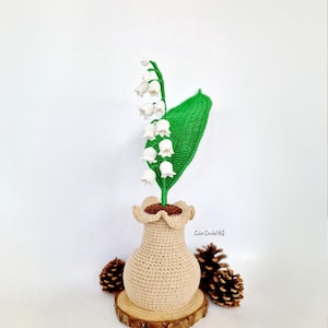 May include: A crocheted lily of the valley arrangement in a beige vase, set on a wooden slice. The arrangement includes white flowers, a green leaf, and brown accents. The vase has a ruffled top. Two pinecones are on either side.
