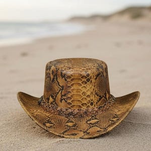 Può includere: Un cappello a tesa larga con un motivo a pelle di serpente marrone e marrone chiaro sulla corona e sulla tesa. Il cappello presenta una fascia in pelle intrecciata attorno alla base della corona, arricchita da piccoli borchie metalliche.