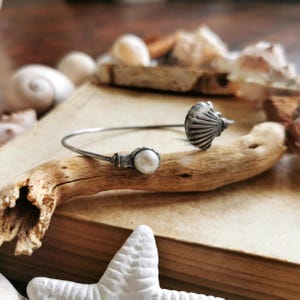 Può includere: Un braccialetto argentato con design a conchiglia e perla. Il braccialetto è esposto su un pezzo di legno trasportato dalle onde, con conchiglie e una stella marina sullo sfondo. Tema nautico.