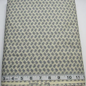 May include: A folded fabric roll with a repeating pattern of small, dark blue floral designs on a cream-colored background. A ruler with inch measurements is visible along the bottom edge. The text "Essentials by Moda" is printed on the fabric.