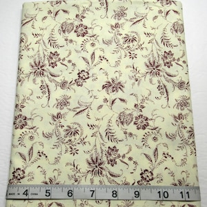 May include: A folded piece of cream-colored fabric featuring a repeating dark red floral and leaf pattern. A white measuring tape is positioned across the bottom edge, showing a width of approximately 11 inches for the fabric piece.