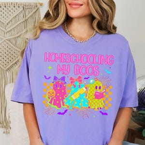 May include: A lilac-colored t-shirt with the text "Homeschooling My Boos" in pink and green. The graphic features three cartoon ghosts with bows, holding a pumpkin, pencil, and book, set against a checkered background with spiderwebs and bats.