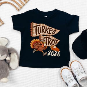 May include: A black short-sleeve t-shirt featuring a graphic of a running turkey wearing a headband and sneakers. The design includes two beige checkered pennants with the text Turkey Trot, a slice of pumpkin pie, and the year 2026 in white.