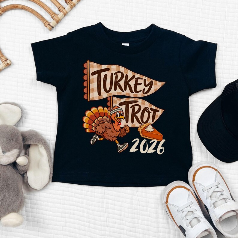 May include: A black short-sleeve t-shirt featuring a graphic of a running turkey wearing a headband and sneakers. The design includes two beige checkered pennants with the text Turkey Trot, a slice of pumpkin pie, and the year 2026 in white.