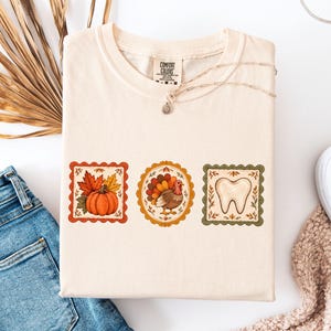 May include: A cream-colored short-sleeve t-shirt featuring three distinct framed illustrations: a pumpkin with autumn leaves, a turkey, and a human tooth. The shirt has a Comfort Colors brand label at the inner neckline and a crew neck collar.