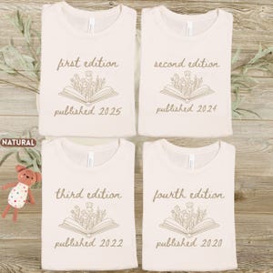 May include: Four cream-colored sweatshirts are displayed, each with a unique design. The designs feature text that reads "first edition," "second edition," "third edition," and "fourth edition," along with publication years and a floral book illustration.