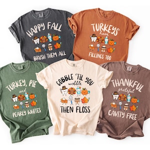 May include: Five short-sleeved t-shirts in charcoal, rust, olive green, cream, and brown. Each shirt features a graphic of a tooth, turkey, toothpaste, pumpkin, pie, spoon, coffee cup, and leaf with text including Happy Fall, Turkeys Have Fillings Too, Turkey Pie and Pearly Whites, Gobble Til You Wobble Then Floss, and Thankful Grateful Cavity Free.