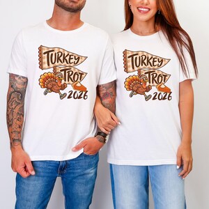 May include: A white t-shirt featuring a graphic of a running turkey wearing a headband and sneakers, accompanied by two checkered pennant flags with the text "TURKEY TROT" and a slice of pumpkin pie, with the year "2026" printed below the design.