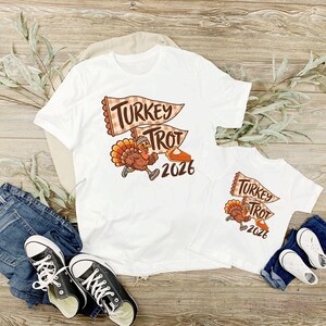 May include: Two white t-shirts featuring a graphic of a cartoon turkey wearing sneakers and a headband, running past a slice of pumpkin pie. Above the turkey are two pennant flags with the text "TURKEY TROT" and the year "2026" below.