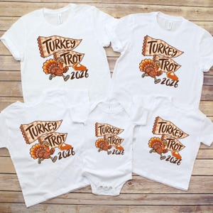 May include: A collection of white t-shirts and a white baby bodysuit featuring a graphic of a running turkey wearing a headband, a checkered pennant flag with the text Turkey Trot, a slice of pumpkin pie, and the year 2026 in brown lettering.