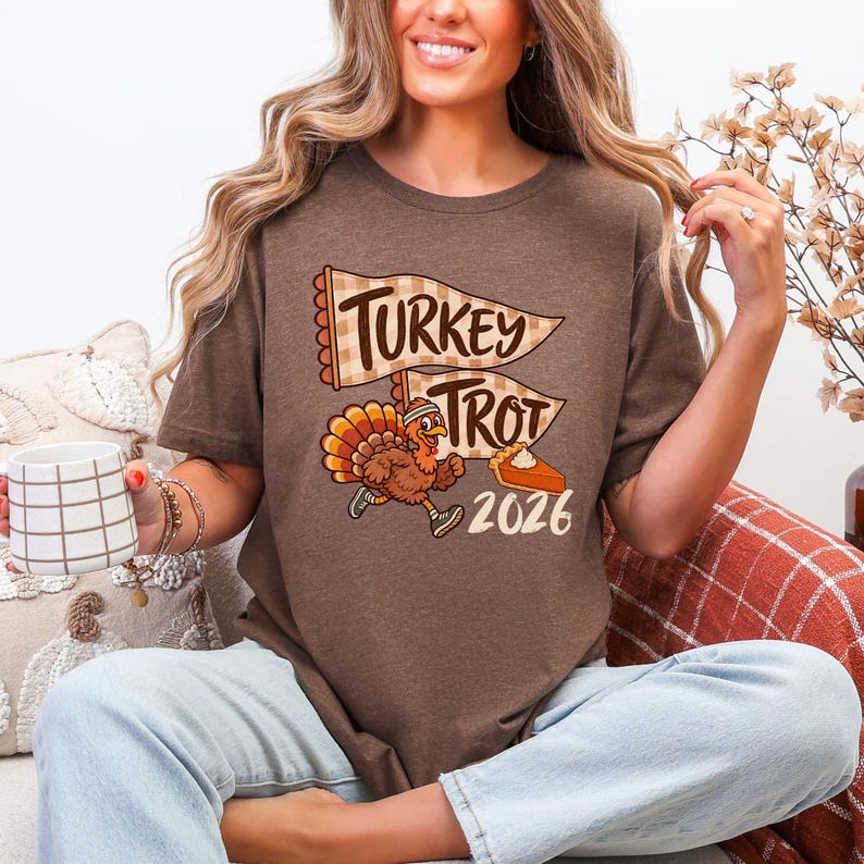 May include: A brown short-sleeve t-shirt featuring a graphic of a running turkey wearing a headband and sneakers. Above the turkey are two checkered pennants with the text "TURKEY TROT" and below is a slice of pumpkin pie and "2026".