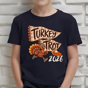 May include: A dark navy blue t-shirt featuring a graphic of a running turkey wearing a headband and sneakers. Above the turkey are two beige checkered pennants with the text "TURKEY TROT" and a slice of pumpkin pie next to the year "2026".